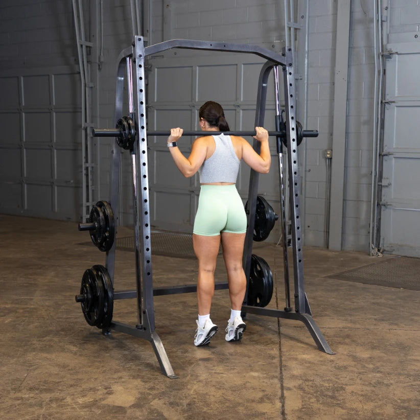 A woman in a gray tank top and light green shorts is preparing to lift a barbell on a squat rack in a gym setting.