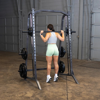 A woman in a gray tank top and light green shorts is preparing to lift a barbell on a squat rack in a gym setting.