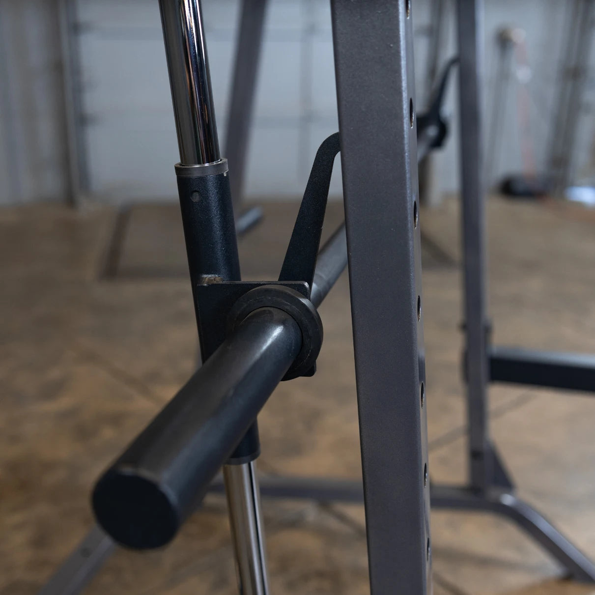 Close-up view of a barbell rack adjustment mechanism, featuring a black lever and chrome pole, set on a concrete floor in a gym environment.