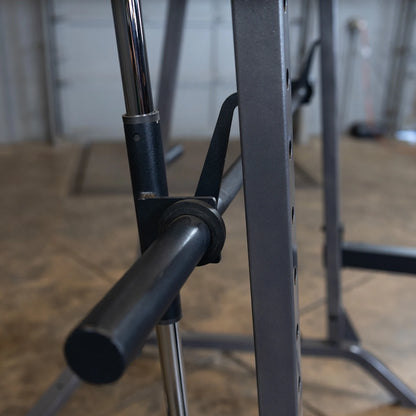 Close-up view of a barbell rack adjustment mechanism, featuring a black lever and chrome pole, set on a concrete floor in a gym environment.