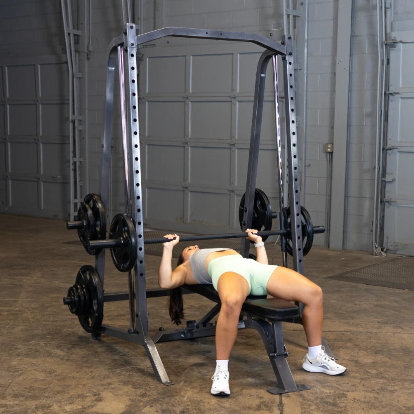 A person performing a bench press exercise in a gym, lying on a bench with a barbell above them, weights on either side, in a spacious workout area.