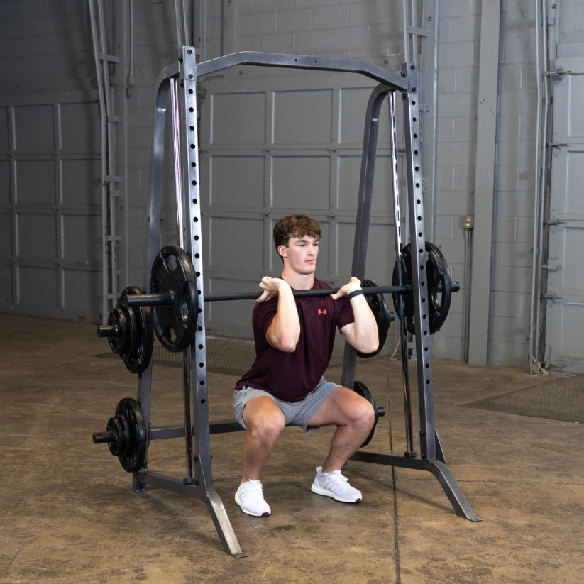 A young man performing a front squat with a barbell in a gym, wearing a maroon shirt and gray shorts, focusing on his form while using a squat rack.