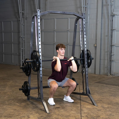 A young man performing a front squat with a barbell in a gym, wearing a maroon shirt and gray shorts, focusing on his form while using a squat rack.