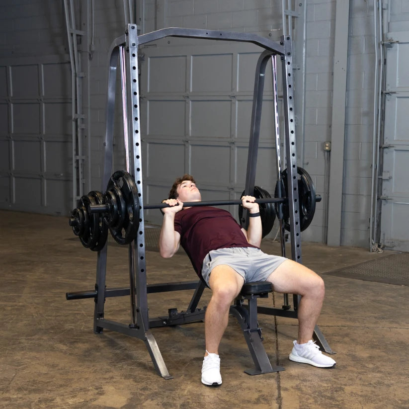 A young man in a maroon shirt and gray shorts is performing a bench press exercise with a barbell loaded with weights, using a squat rack in a gym setting.