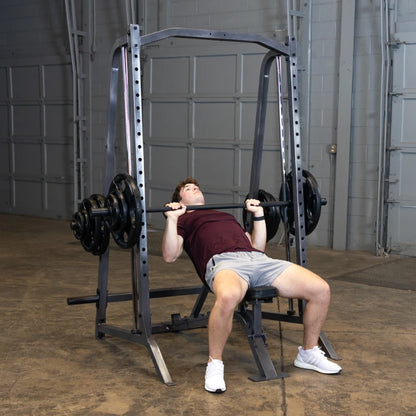 A young man in a maroon shirt and gray shorts is performing a bench press exercise with a barbell loaded with weights, using a squat rack in a gym setting.