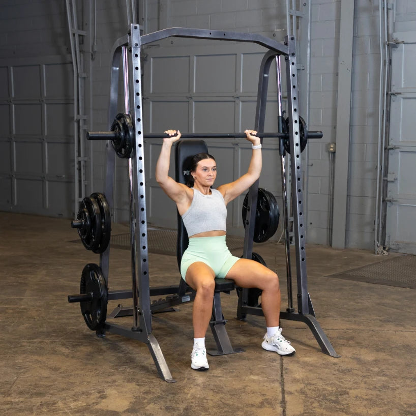 A woman performing an overhead press exercise in a gym, seated on a weightlifting machine with a barbell above her head, wearing a gray crop top and mint green shorts.
