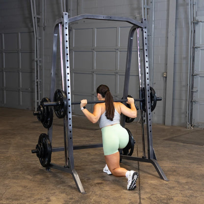 A woman in a gray sports bra and mint green shorts performing a squat with a barbell on a squat rack in a gym setting.