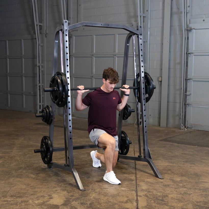 A young man performing a barbell squat exercise inside a gym, with a squat rack and weights in the background.