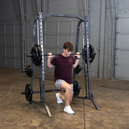 A young man performing a barbell squat exercise inside a gym, with a squat rack and weights in the background.