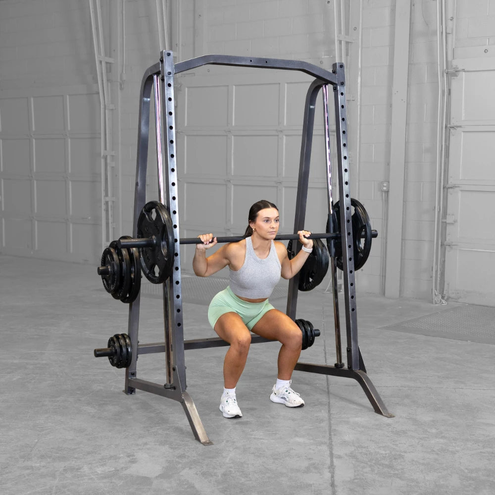 A woman in a gray tank top and green shorts performs a squat with a barbell on a squat rack in a gym setting.