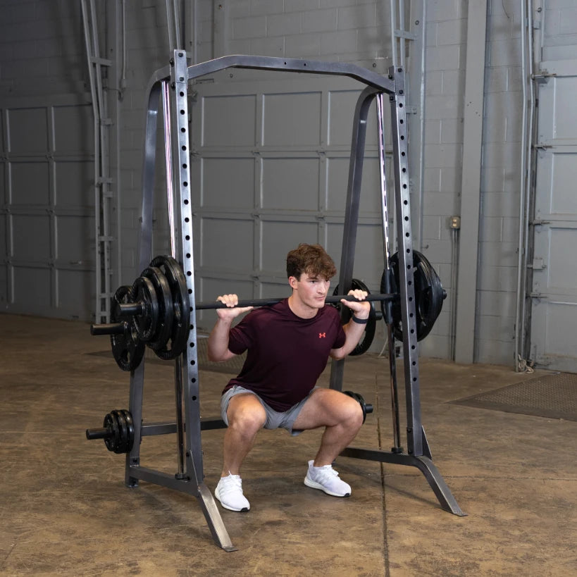 A young man performing a squat exercise with a barbell on a squat rack in a gym setting. He is wearing a maroon t-shirt and shorts, focusing on his form while lifting weights.