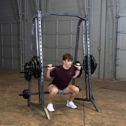 A young man performing a squat exercise with a barbell on a squat rack in a gym setting. He is wearing a maroon t-shirt and shorts, focusing on his form while lifting weights.