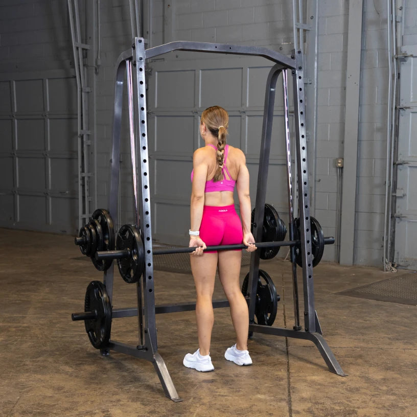 A woman in a pink sports bra and shorts performs a deadlift using a barbell in a gym setting, with a weight rack in the background.
