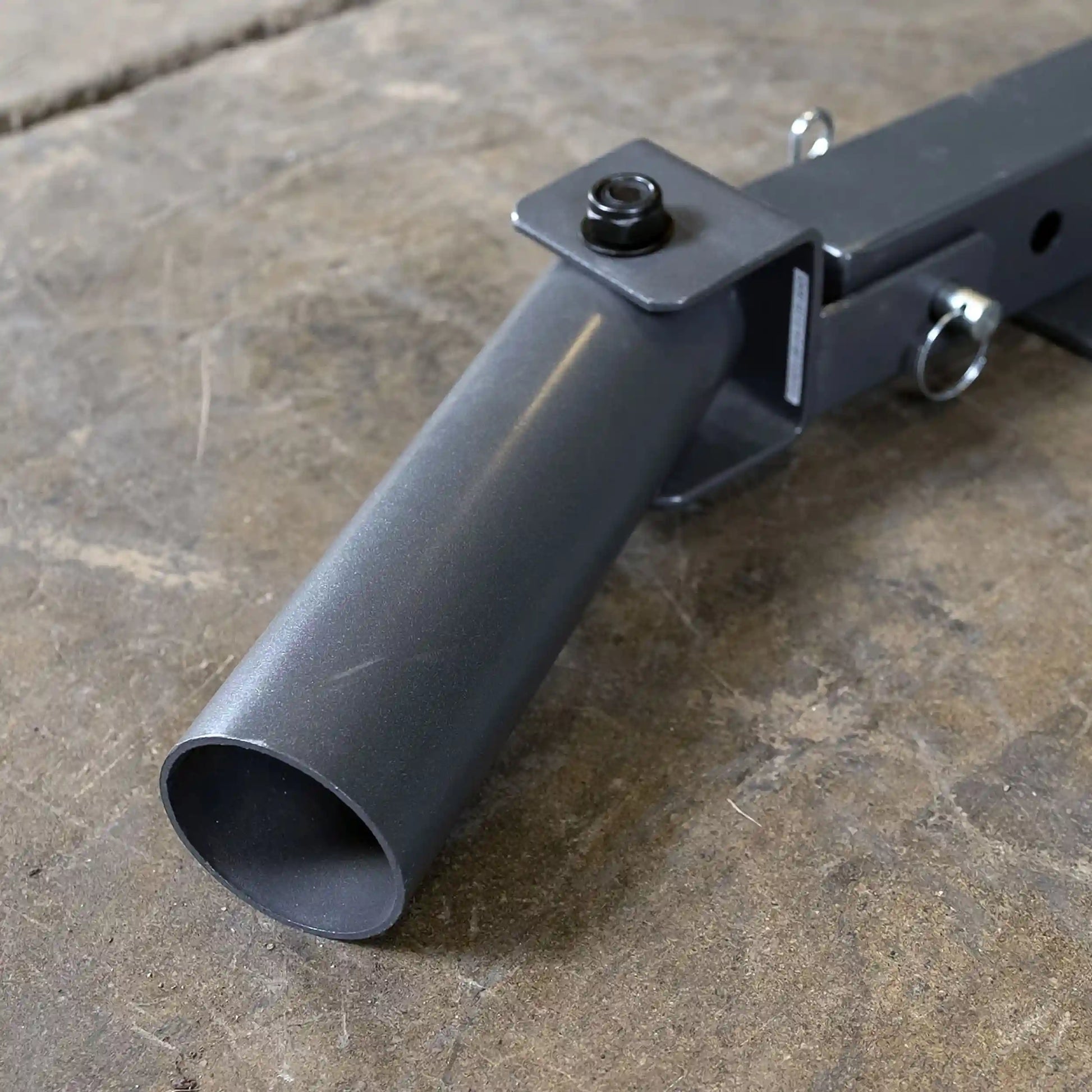 Close-up of a gray metal pipe attachment with a square mounting bracket and a bolt, resting on a textured concrete surface.