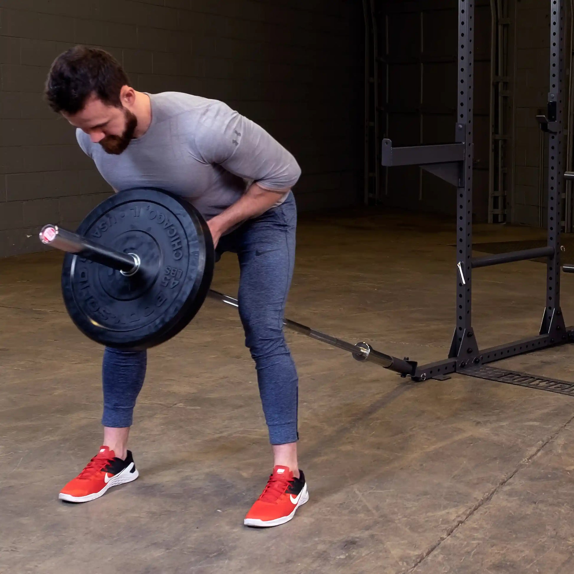 A man with a beard wearing a gray long-sleeve shirt and blue athletic pants is performing a bent-over row exercise with a barbell in a gym setting. He is wearing red and black athletic shoes, and a squat rack is visible in the background.