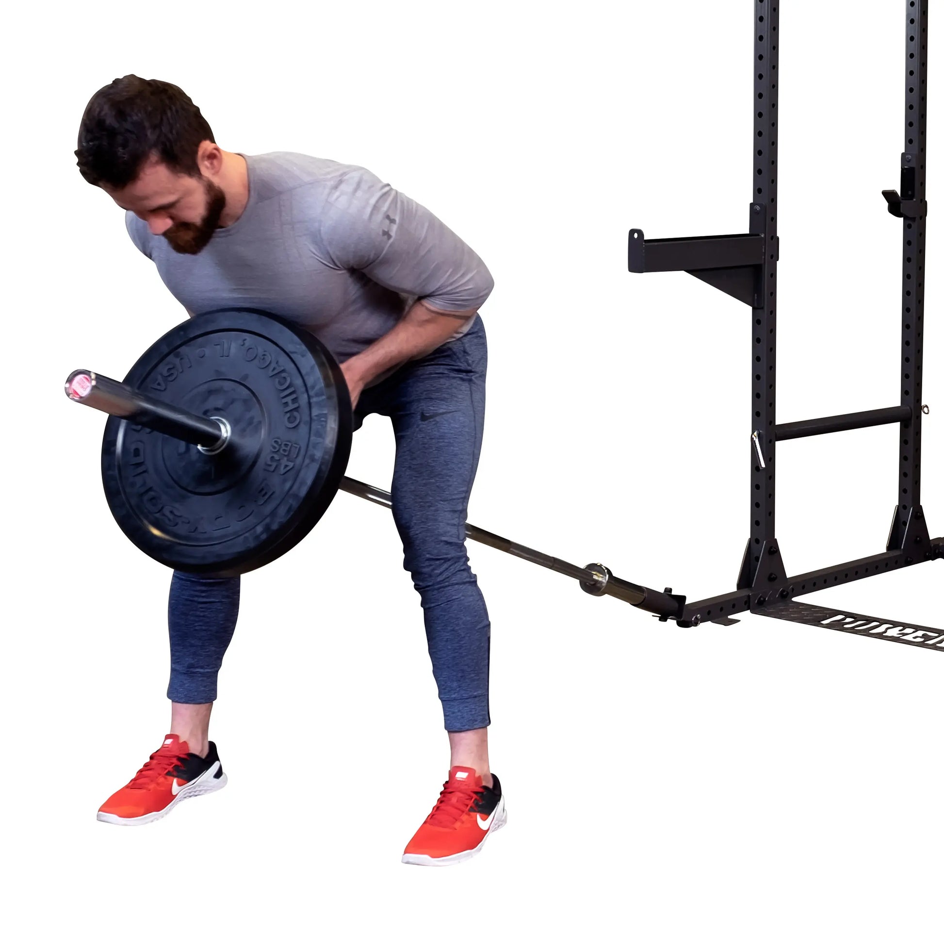A man in a gray shirt and blue athletic pants performs a bent-over row with a barbell in a gym setting. He is focused on lifting the weight, which has black plates on either side, while wearing red and black athletic shoes. A squat rack is visible in the background.
