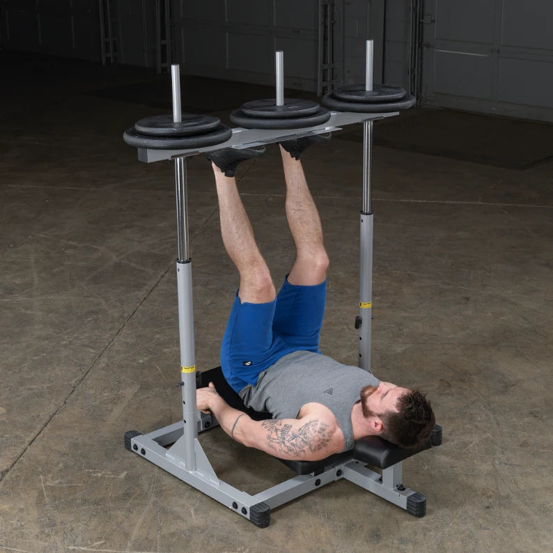 A man performing a leg exercise on a weight bench, with his legs elevated and holding onto the bench. He is wearing a gray tank top and blue shorts, and there are weight plates on the bar above him. The setting is a gym with a concrete floor.