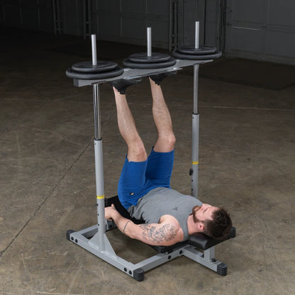 A man performing a leg exercise on a weight bench, with his legs elevated and holding onto the bench. He is wearing a gray tank top and blue shorts, and there are weight plates on the bar above him. The setting is a gym with a concrete floor.
