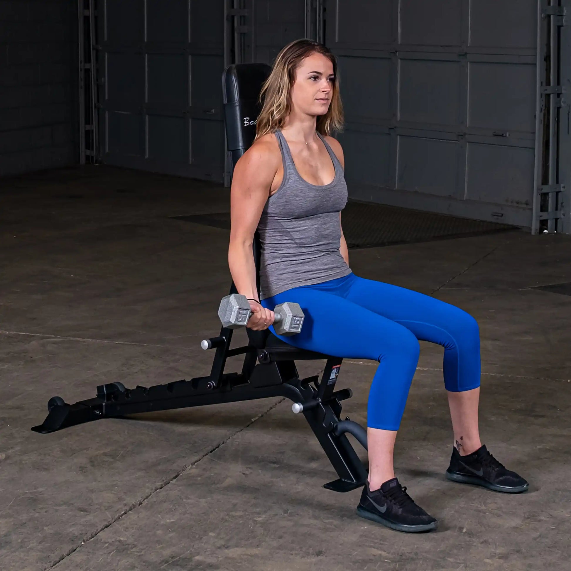 A woman sitting on the Body-Solid Pro ClubLine Adjustable Bench SFID325, holding dumbbells in each hand, wearing a gray tank top and blue leggings, in a gym setting.