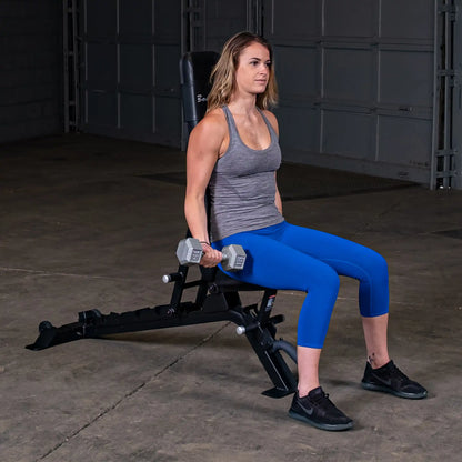 A woman sitting on the Body-Solid Pro ClubLine Adjustable Bench SFID325, holding dumbbells in each hand, wearing a gray tank top and blue leggings, in a gym setting.