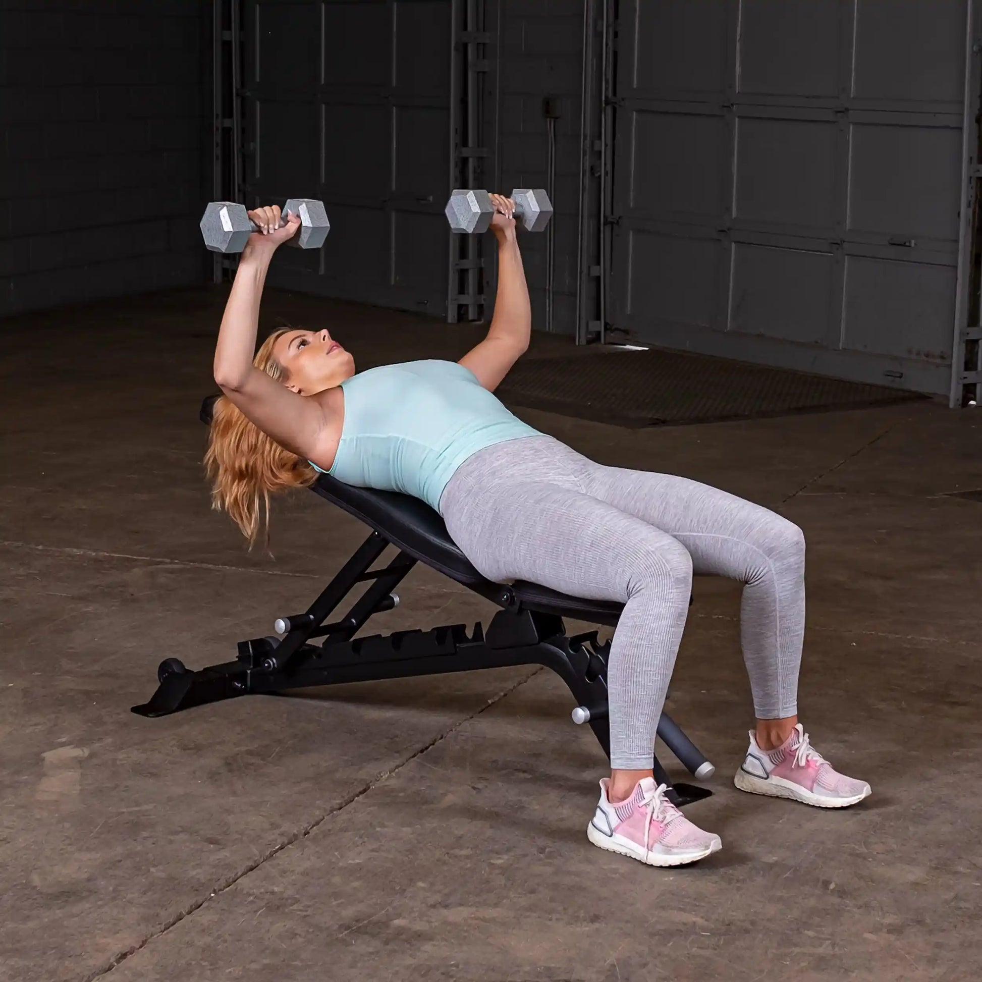 A woman lying on a Body-Solid Pro ClubLine Adjustable Bench SFID325, performing a dumbbell press with weights in both hands in a gym setting.