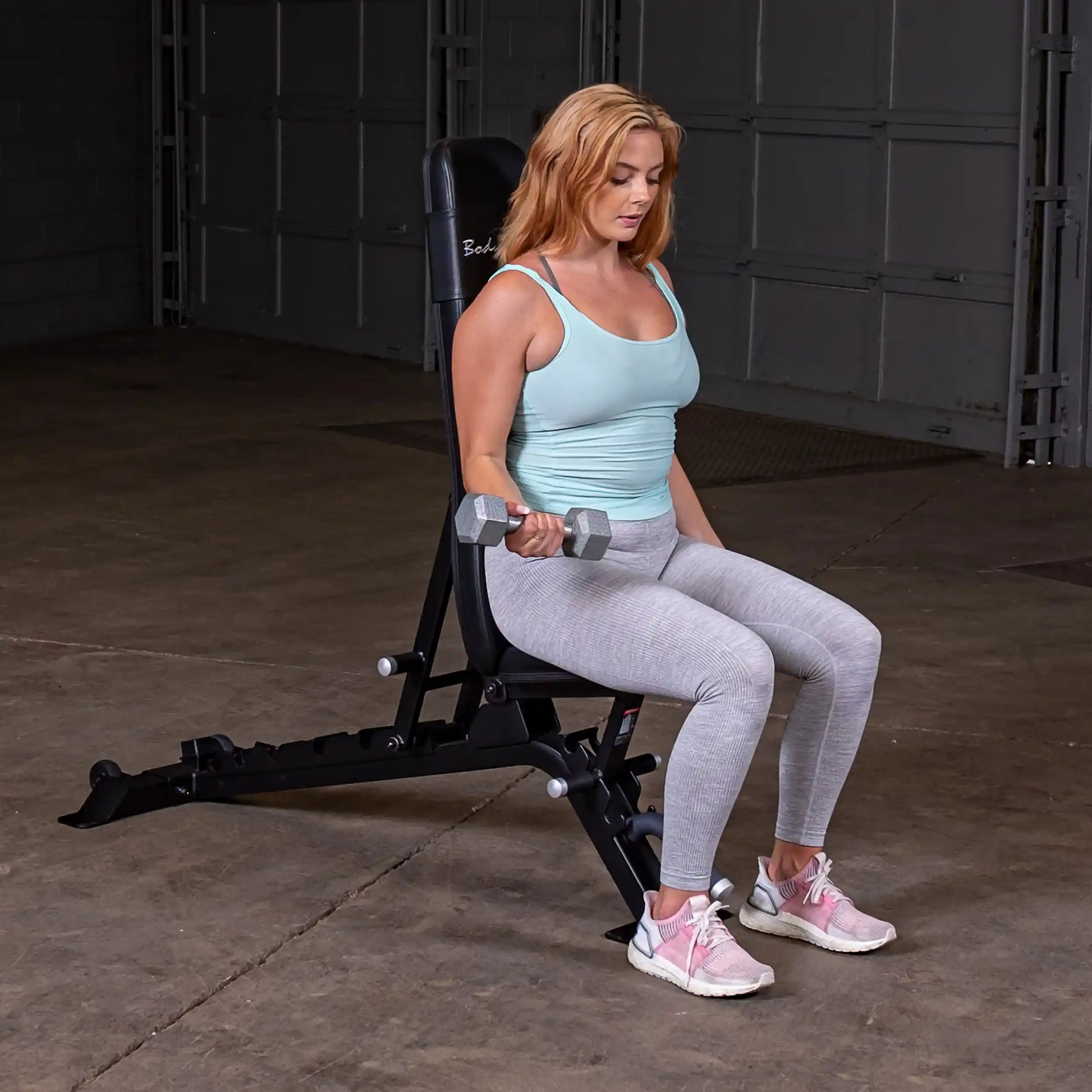 A woman sitting on the Body-Solid Pro ClubLine Adjustable Bench SFID325, performing a dumbbell exercise with a focused expression, in a gym setting.