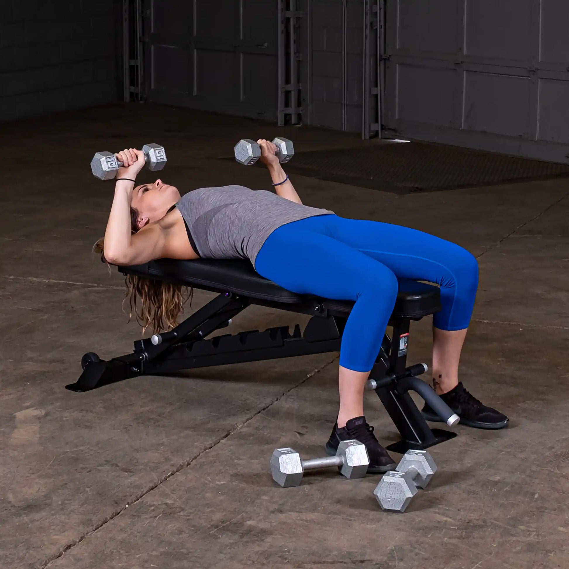 A woman lying on the Body-Solid Pro ClubLine Adjustable Bench SFID325, performing an exercise with dumbbells in each hand, wearing a gray tank top and blue leggings in a gym setting.