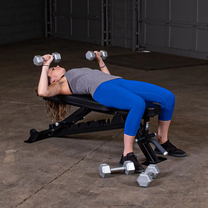 A woman lying on the Body-Solid Pro ClubLine Adjustable Bench SFID325, performing an exercise with dumbbells in each hand, wearing a gray tank top and blue leggings in a gym setting.