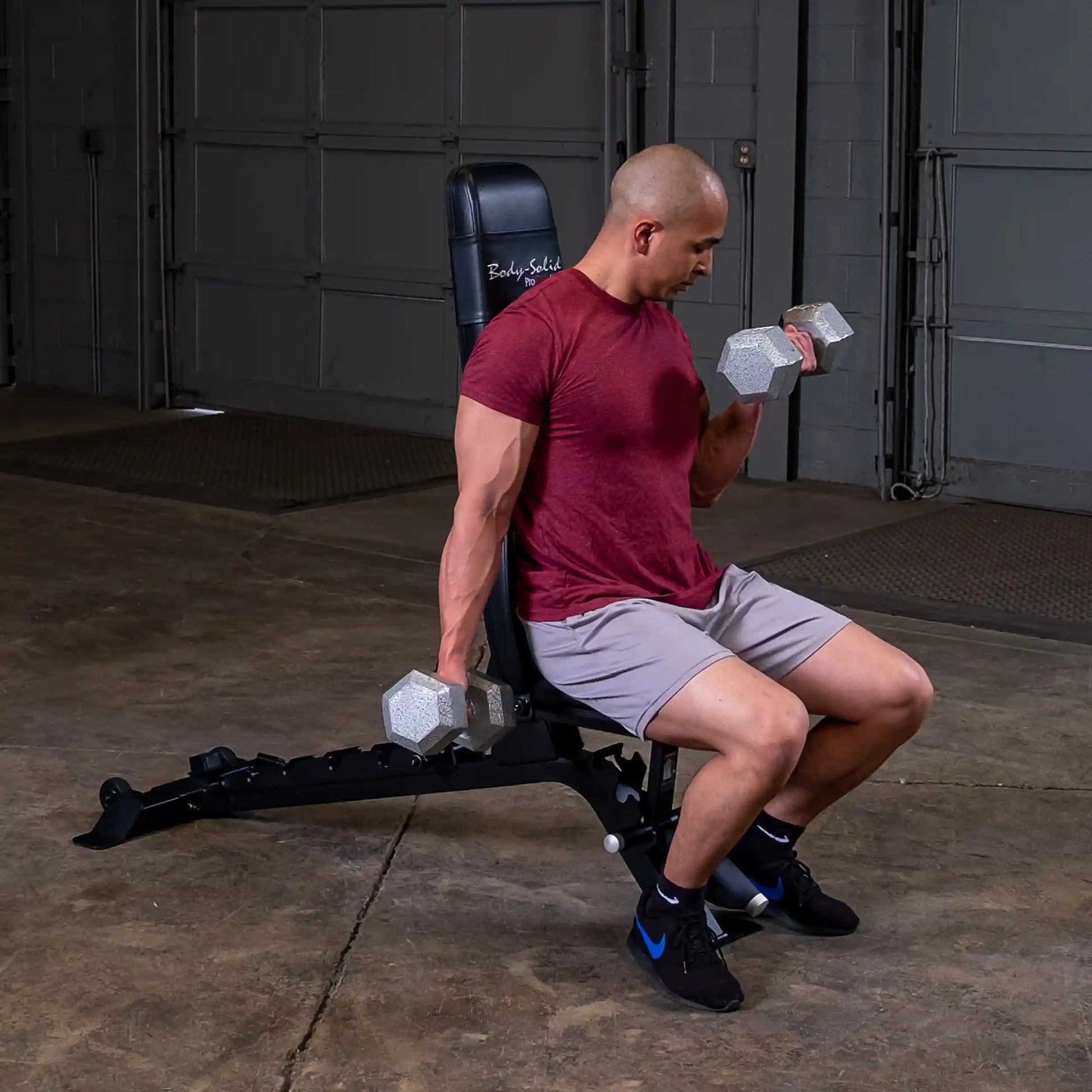 A man sits on a Body-Solid Pro ClubLine Adjustable Bench SFID325, performing a dumbbell curl with two hexagonal dumbbells in a gym setting.
