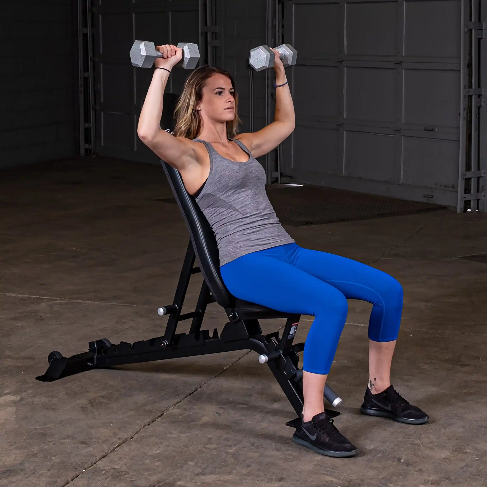 A woman sitting on the Body-Solid Pro ClubLine Adjustable Bench SFID325, performing an overhead dumbbell press with two hexagonal weights in a gym setting.