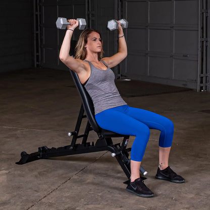 A woman sitting on the Body-Solid Pro ClubLine Adjustable Bench SFID325, performing an overhead dumbbell press with two hexagonal weights in a gym setting.