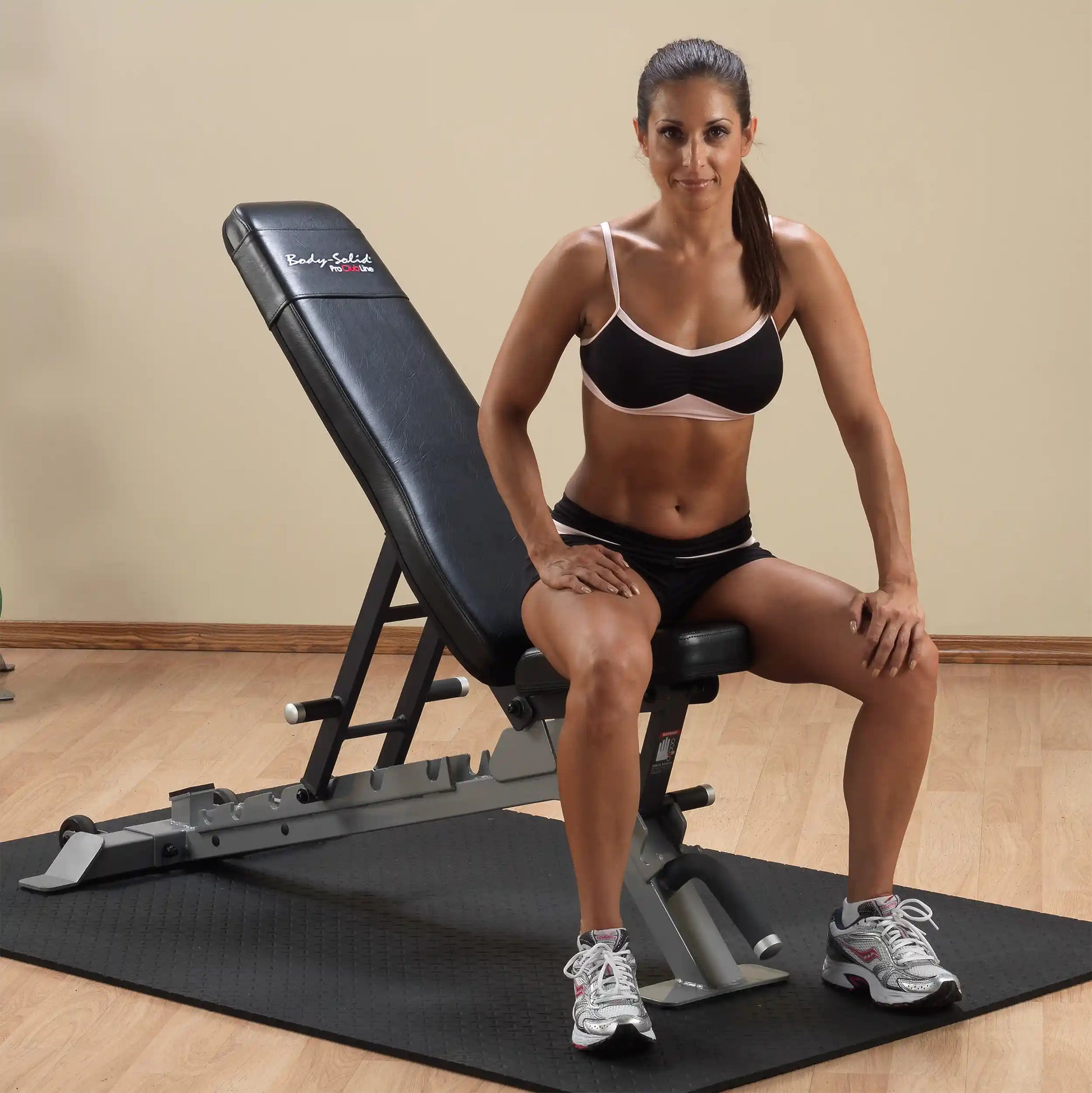 A fit woman sitting on the Body-Solid Pro ClubLine Adjustable Bench SFID325, wearing athletic attire, with a neutral background and gym flooring.