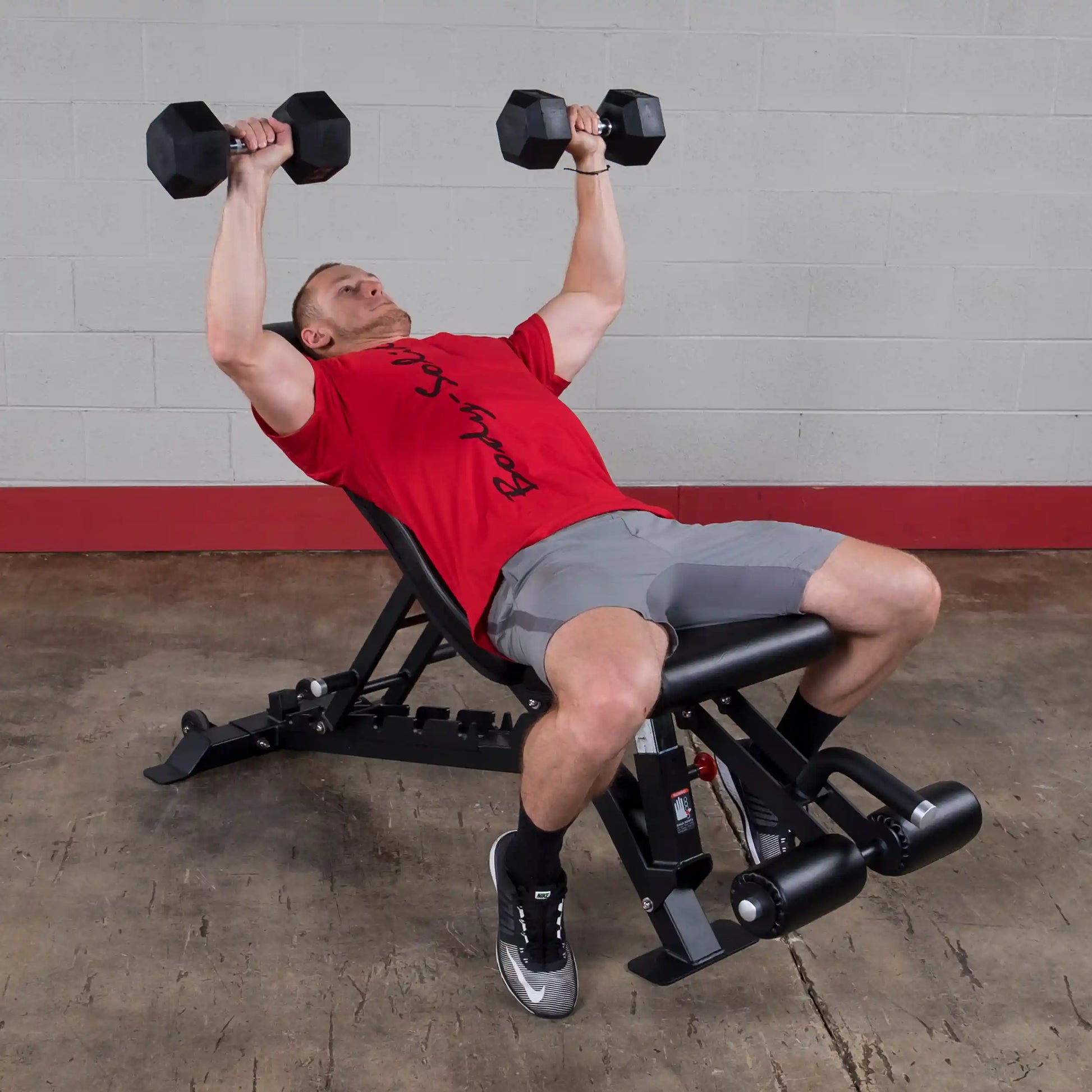 A person exercising on a Body-Solid Pro ClubLine Adjustable Bench SFID425, performing a dumbbell press with weights in both hands, wearing a red shirt and gray shorts.
