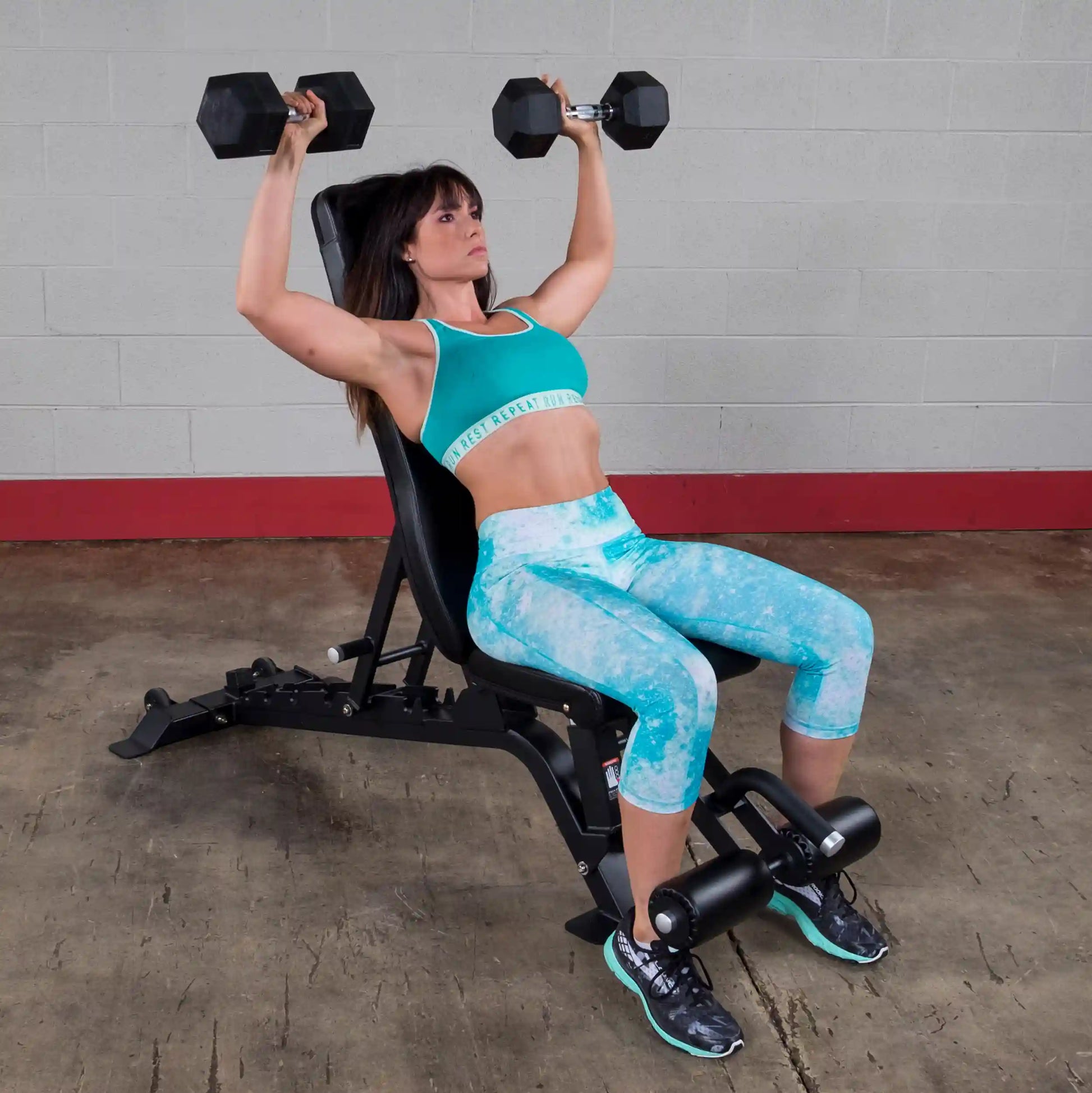 A woman performing a dumbbell shoulder press on the Body-Solid Pro ClubLine Adjustable Bench SFID425, wearing a turquoise sports bra and matching leggings.
