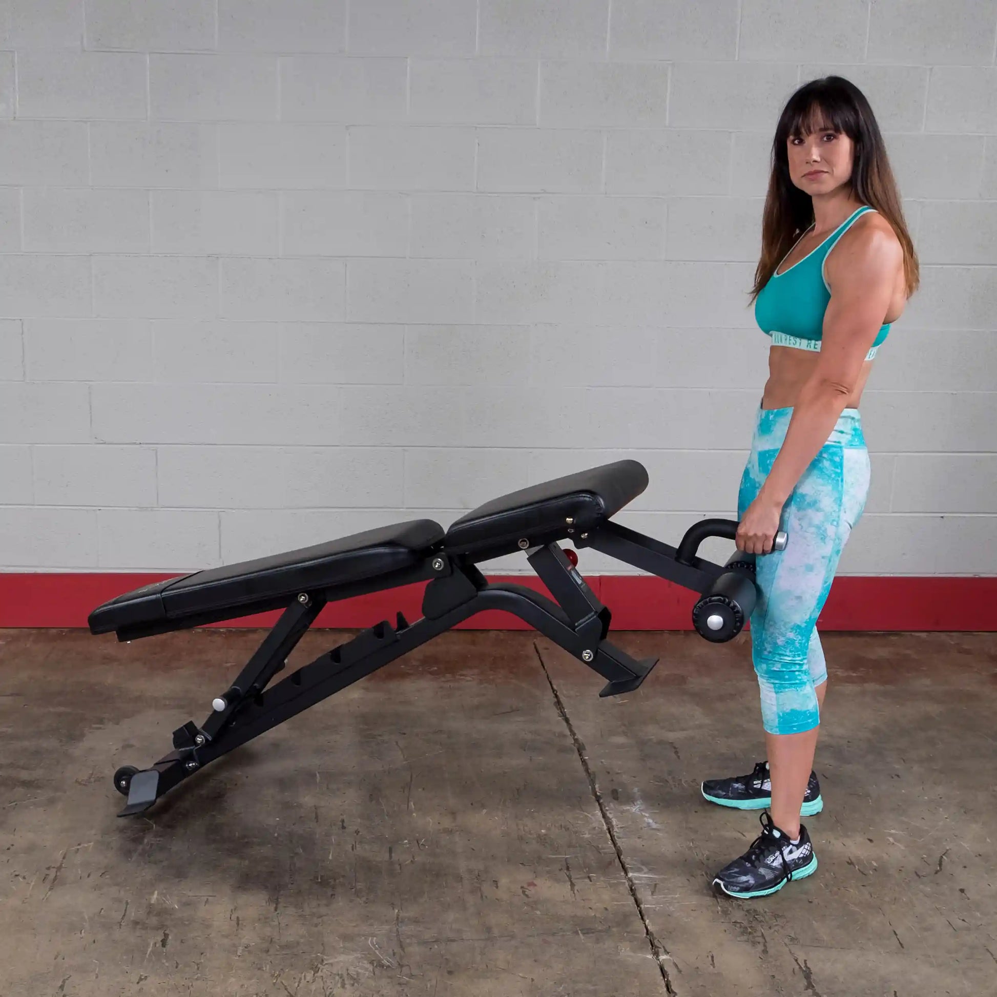 A woman in athletic wear lifting the Body-Solid Pro ClubLine Adjustable Bench SFID425 in a gym setting with a plain wall in the background.