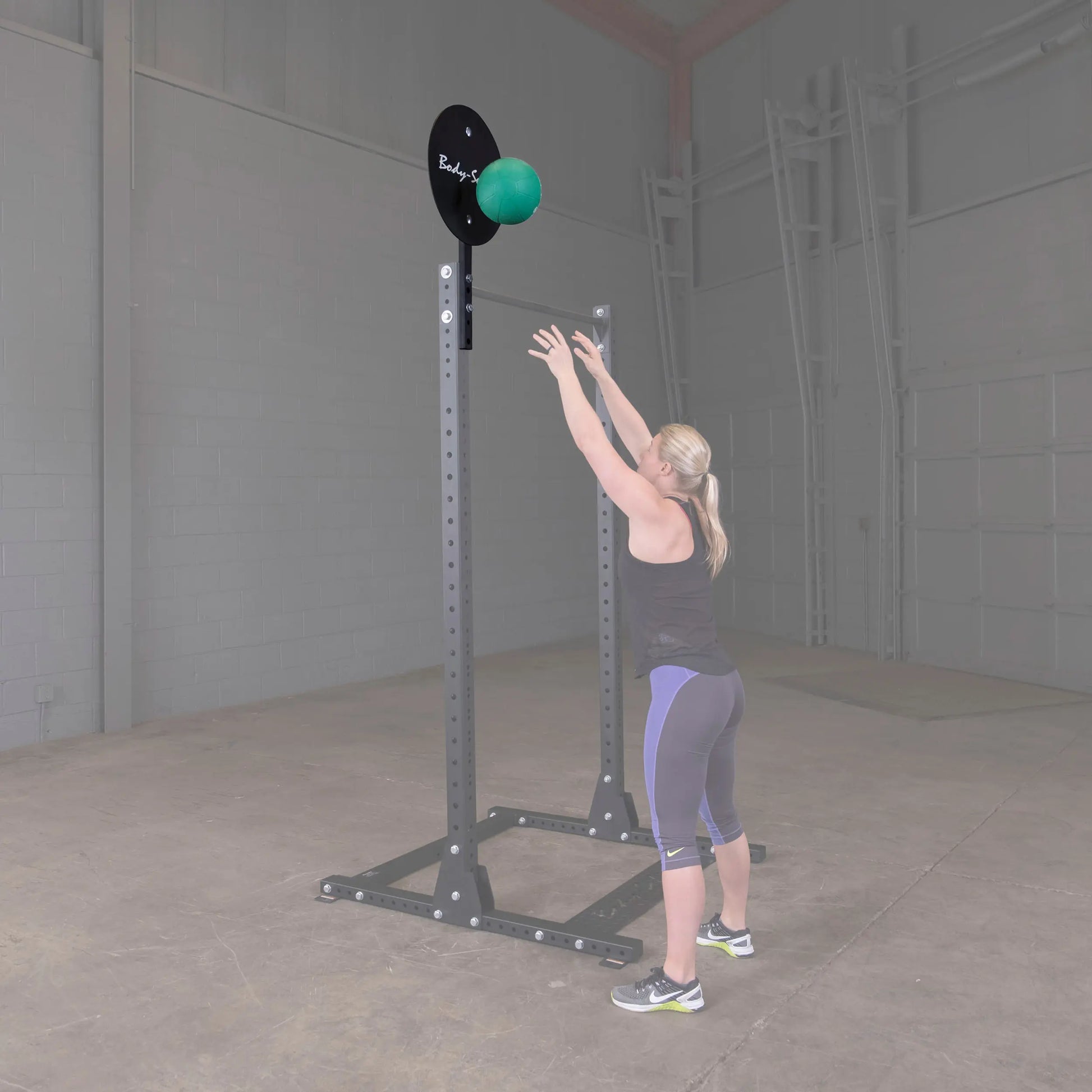 A woman is exercising with the Body-Solid Pro ClubLine Ball Target Attachment SPRBT in a gym setting, reaching up towards a green medicine ball attached to a black target.