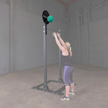 A woman is exercising with the Body-Solid Pro ClubLine Ball Target Attachment SPRBT in a gym setting, reaching up towards a green medicine ball attached to a black target.