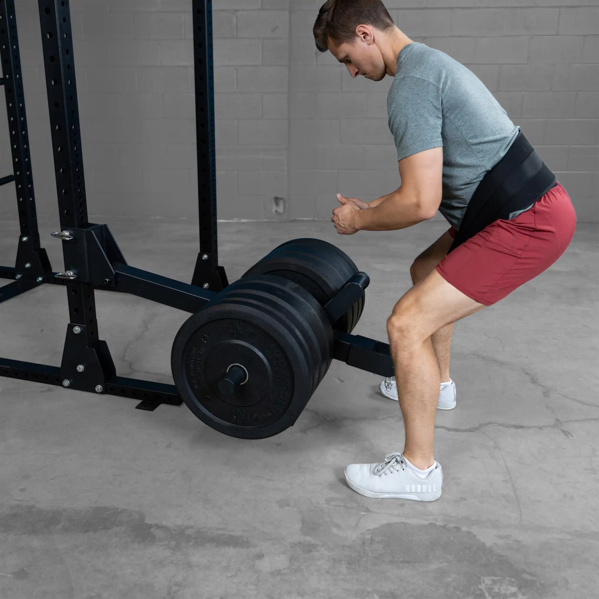 A person performing a squat using the Body-Solid Pro ClubLine Belt Squat Attachment SPRBSA, featuring a barbell loaded with weights, in a gym setting with a concrete floor and gray backdrop.