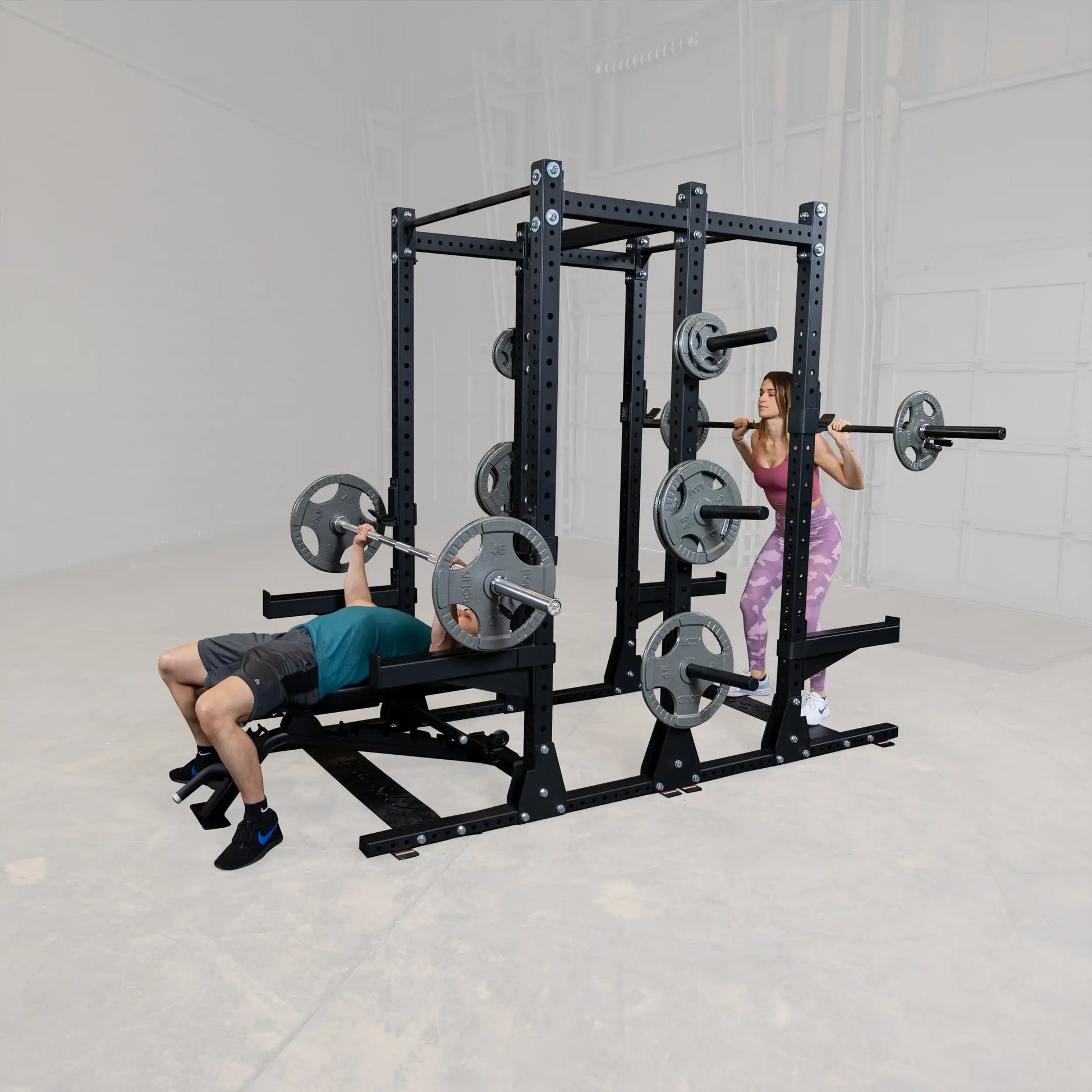 Two individuals using the Body-Solid Pro ClubLine Commercial Double Half Rack Kit SPRDBLKIT; one person is performing a bench press while the other is doing a squat, showcasing the equipment's versatility in a gym setting.
