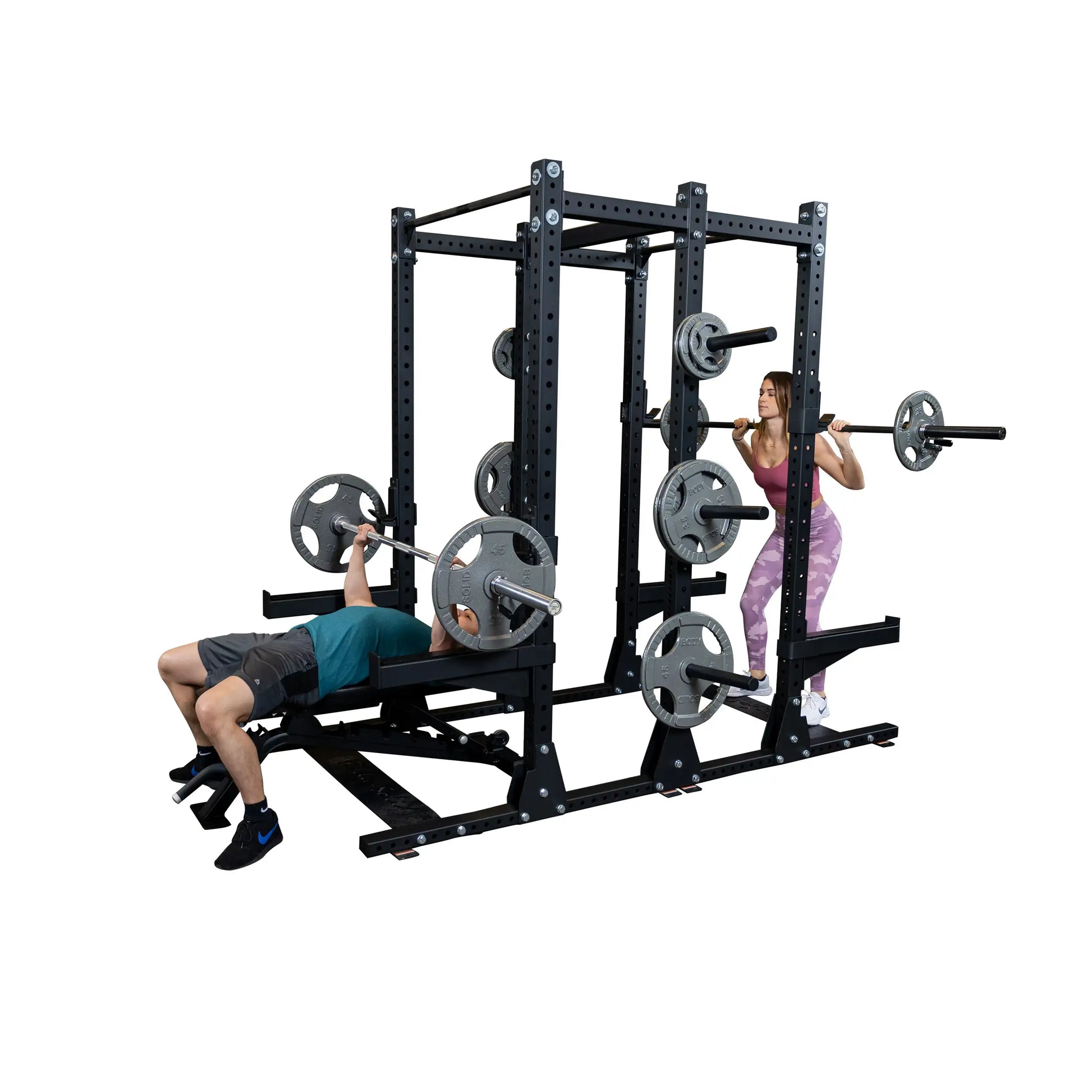 Two people exercising with the Body-Solid Pro ClubLine Commercial Double Half Rack Kit SPRDBLKIT. One person is performing a bench press, while the other is doing a squat, both using the rack for support.