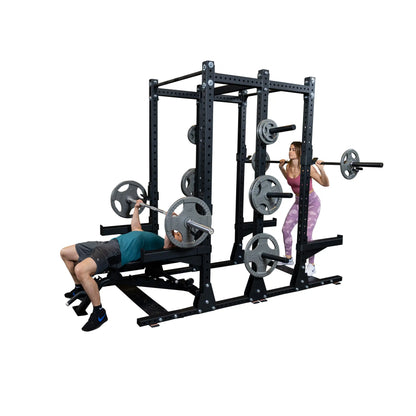 Two people exercising with the Body-Solid Pro ClubLine Commercial Double Half Rack Kit SPRDBLKIT. One person is performing a bench press, while the other is doing a squat, both using the rack for support.