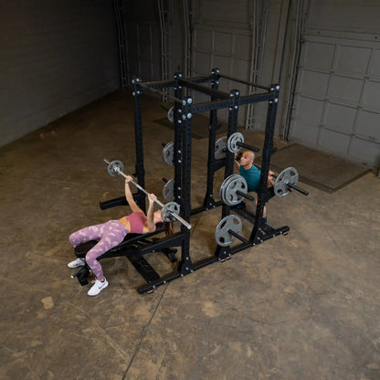 Two people working out in a gym using the Body-Solid Pro ClubLine Commercial Double Half Rack Kit SPRDBLKIT, performing exercises with barbells and weights on the rack.