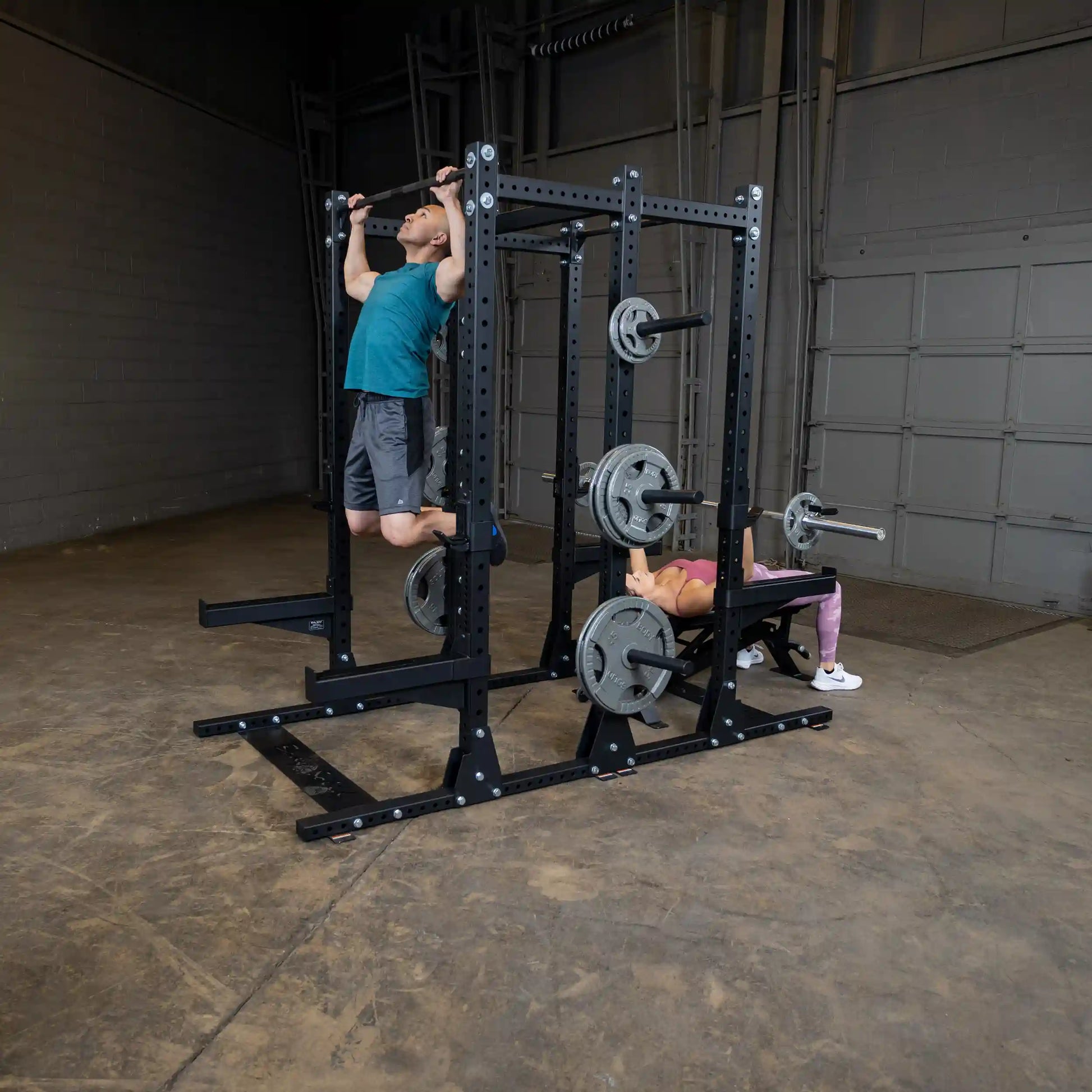 An image showing two individuals exercising on a Body-Solid Pro ClubLine Commercial Double Half Rack Kit SPRDBLKIT. One person is performing pull-ups, while the other is doing bench presses in a gym setting.