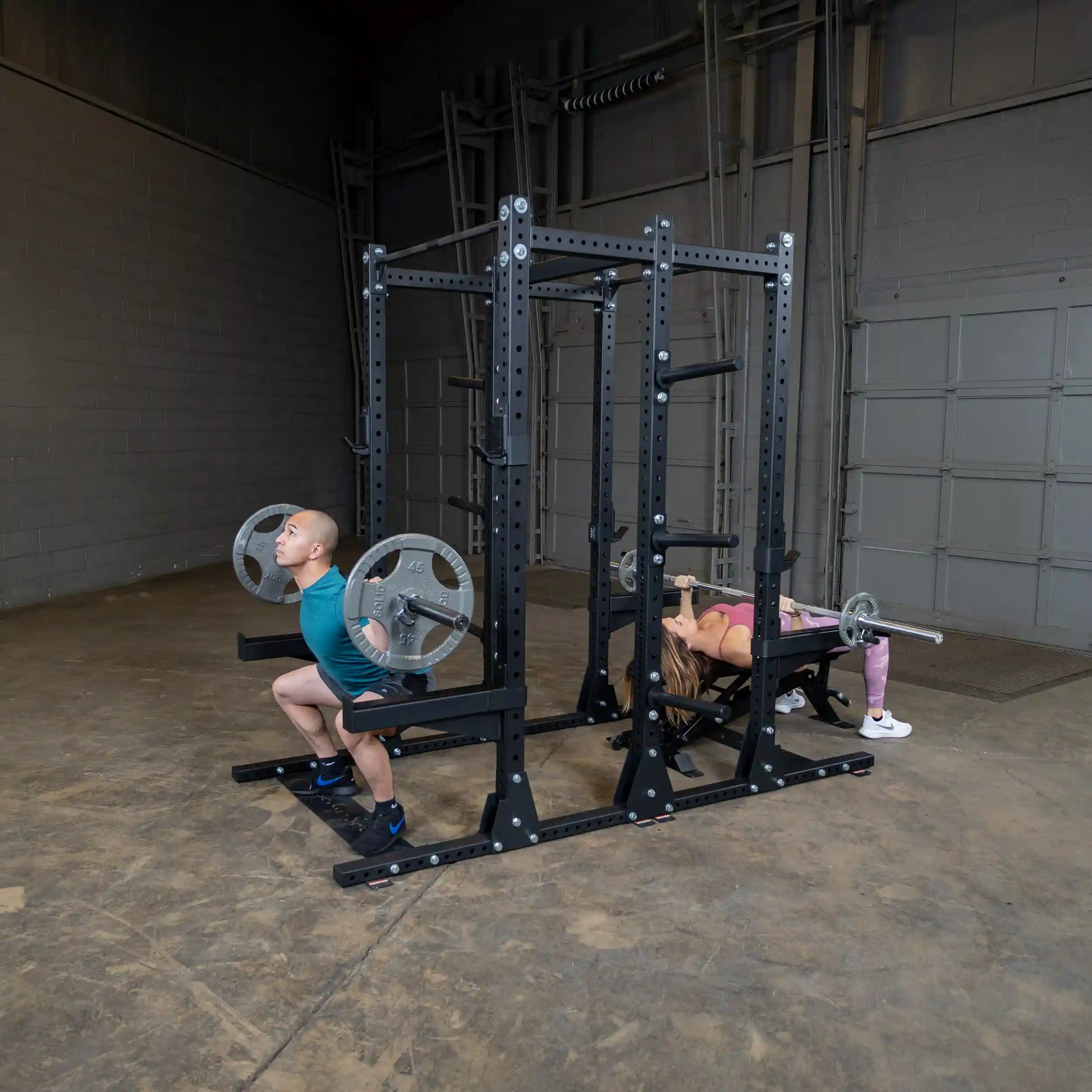 Two individuals exercising on the Body-Solid Pro ClubLine Commercial Double Half Rack Kit SPRDBLKIT, one performing a squat and the other doing a bench press.