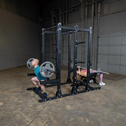 Two individuals exercising on the Body-Solid Pro ClubLine Commercial Double Half Rack Kit SPRDBLKIT, one performing a squat and the other doing a bench press.
