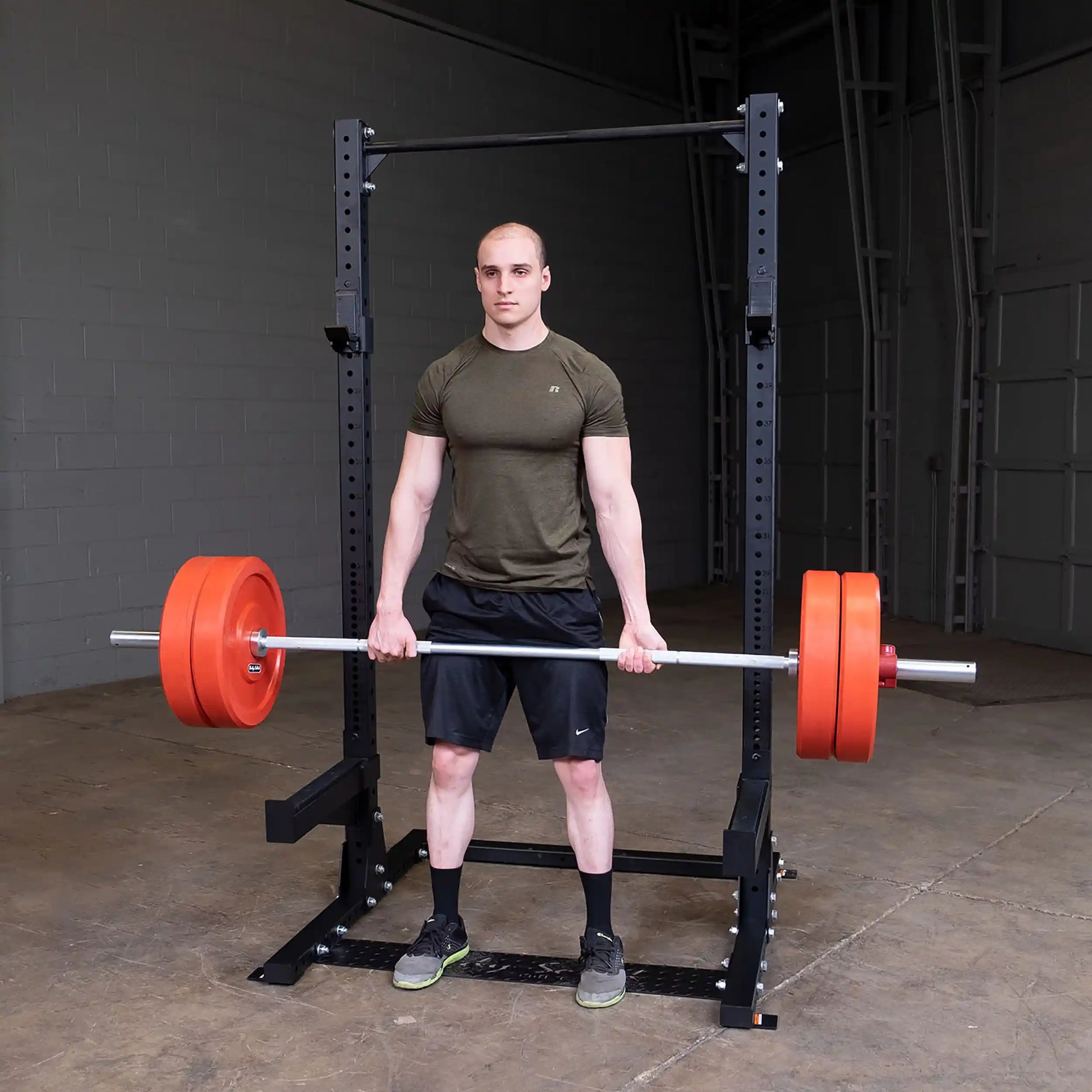 A person performing a deadlift with a barbell loaded with orange weights, standing in front of a Body-Solid Pro ClubLine Commercial Half Rack SPR500 in a gym setting.