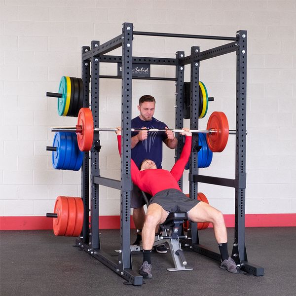 Two individuals are using the Body-Solid Pro ClubLine Commercial Power Rack Extension SPRBACK. One person is bench pressing with weights, while another spots them inside a sturdy black power rack, with various colored weight plates mounted on the sides.