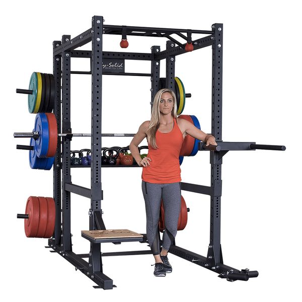 A woman in athletic wear is standing next to a Body-Solid Pro ClubLine Commercial Power Rack Extension SPRBACK, which is loaded with colorful weight plates and kettlebells.