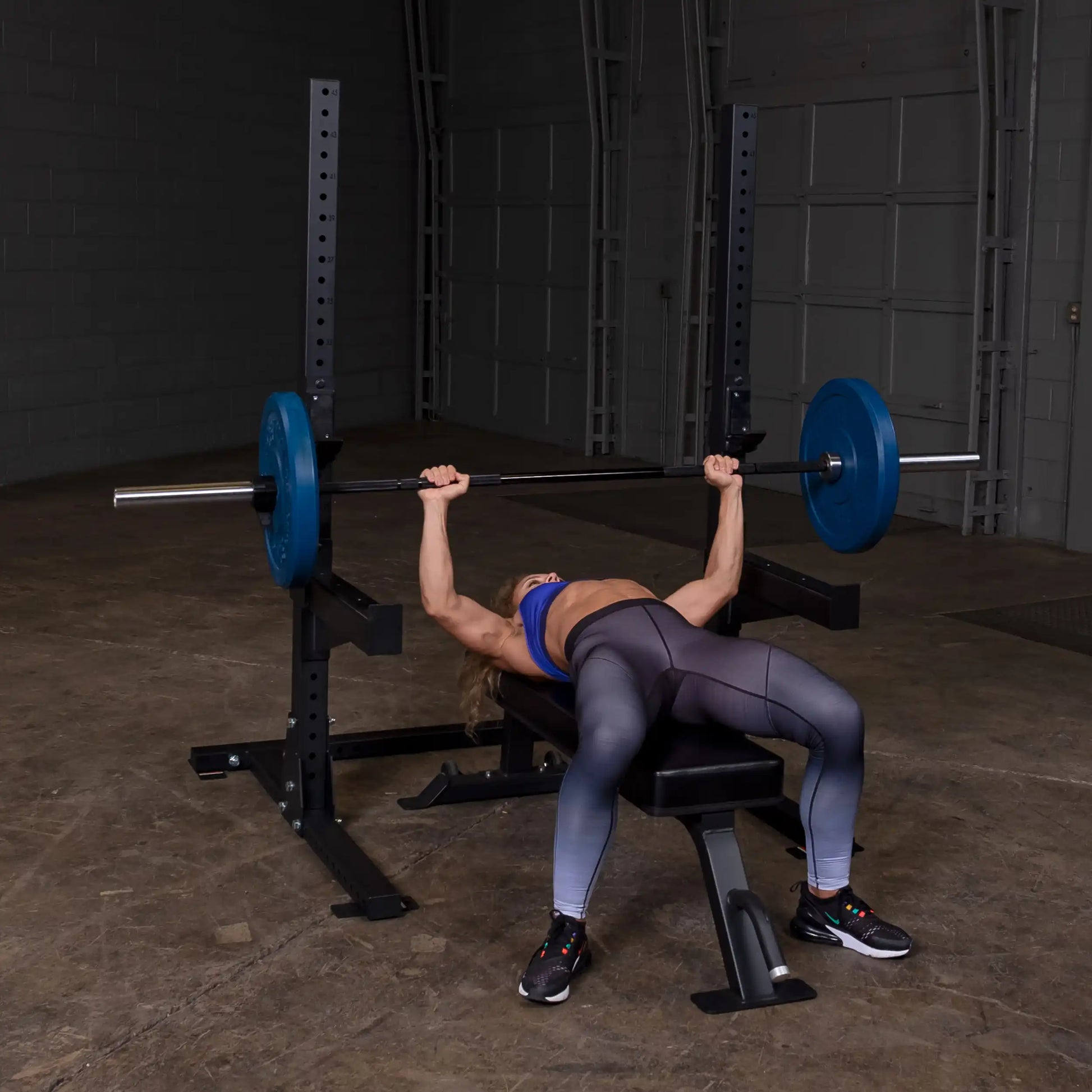 A person performing a bench press in a gym, using a barbell with blue weight plates on a Body-Solid Pro ClubLine Commercial Squat Stand SPR250.