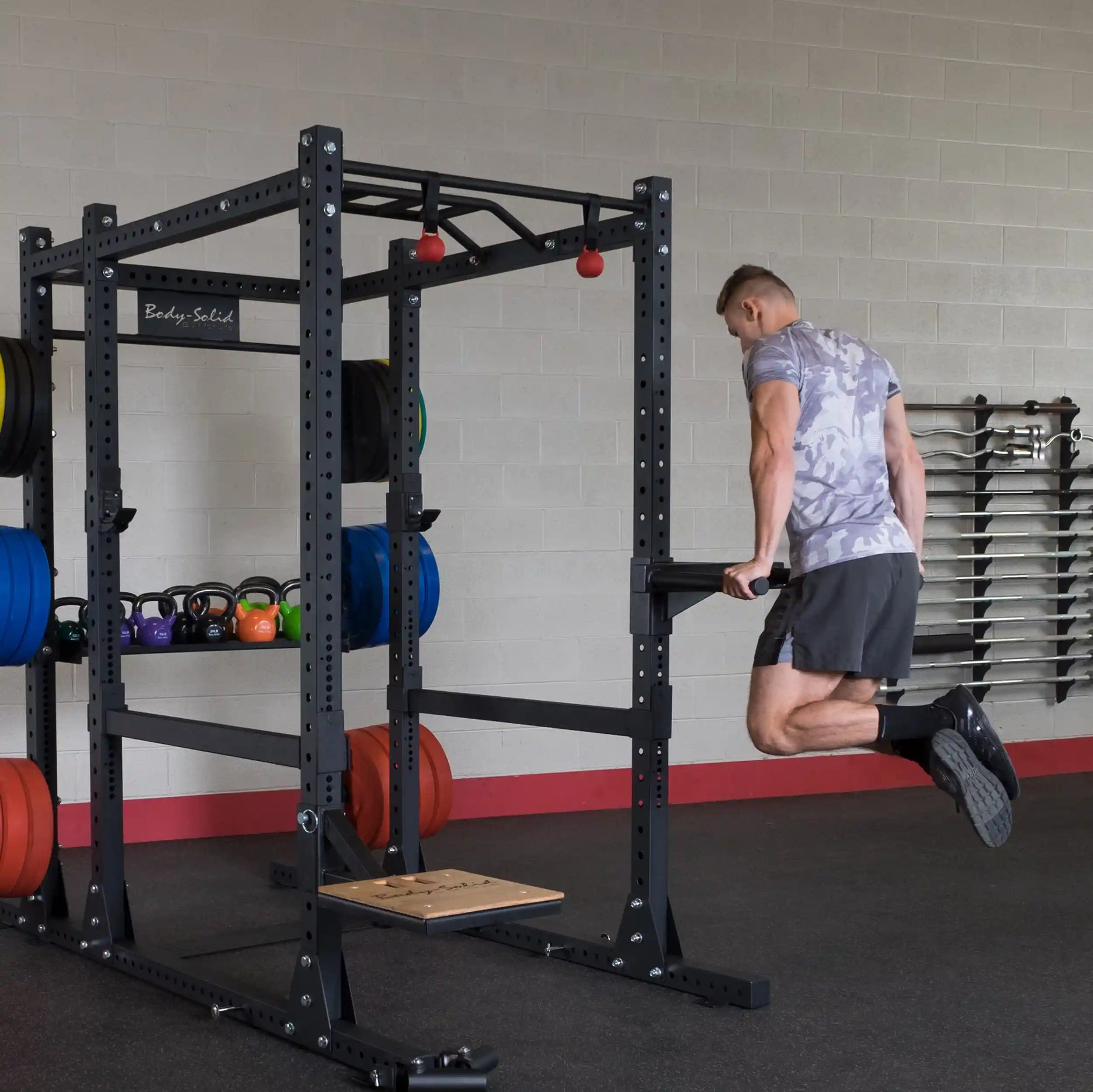 A person performing a dip exercise using the Body-Solid Pro ClubLine Dip Station Attachment SPRDIP, with colorful weight plates and kettlebells in the background.